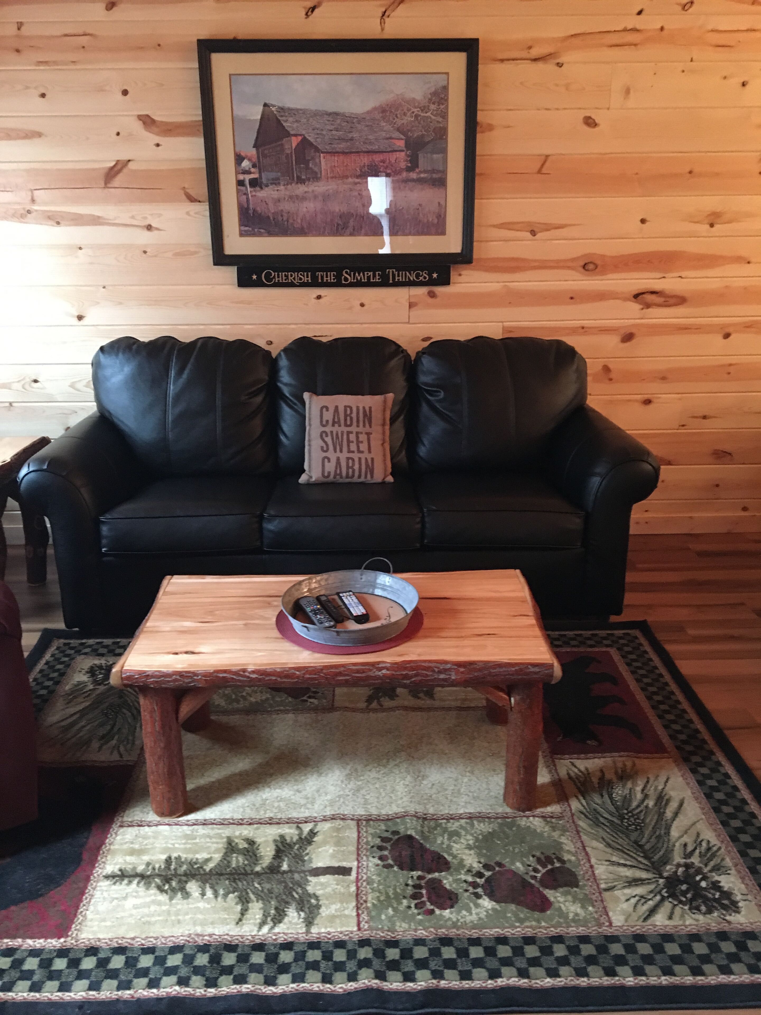 A cozy living room with a black leather sofa, a wooden coffee table with remotes, a framed picture on the wall, and a rug with a forest design.