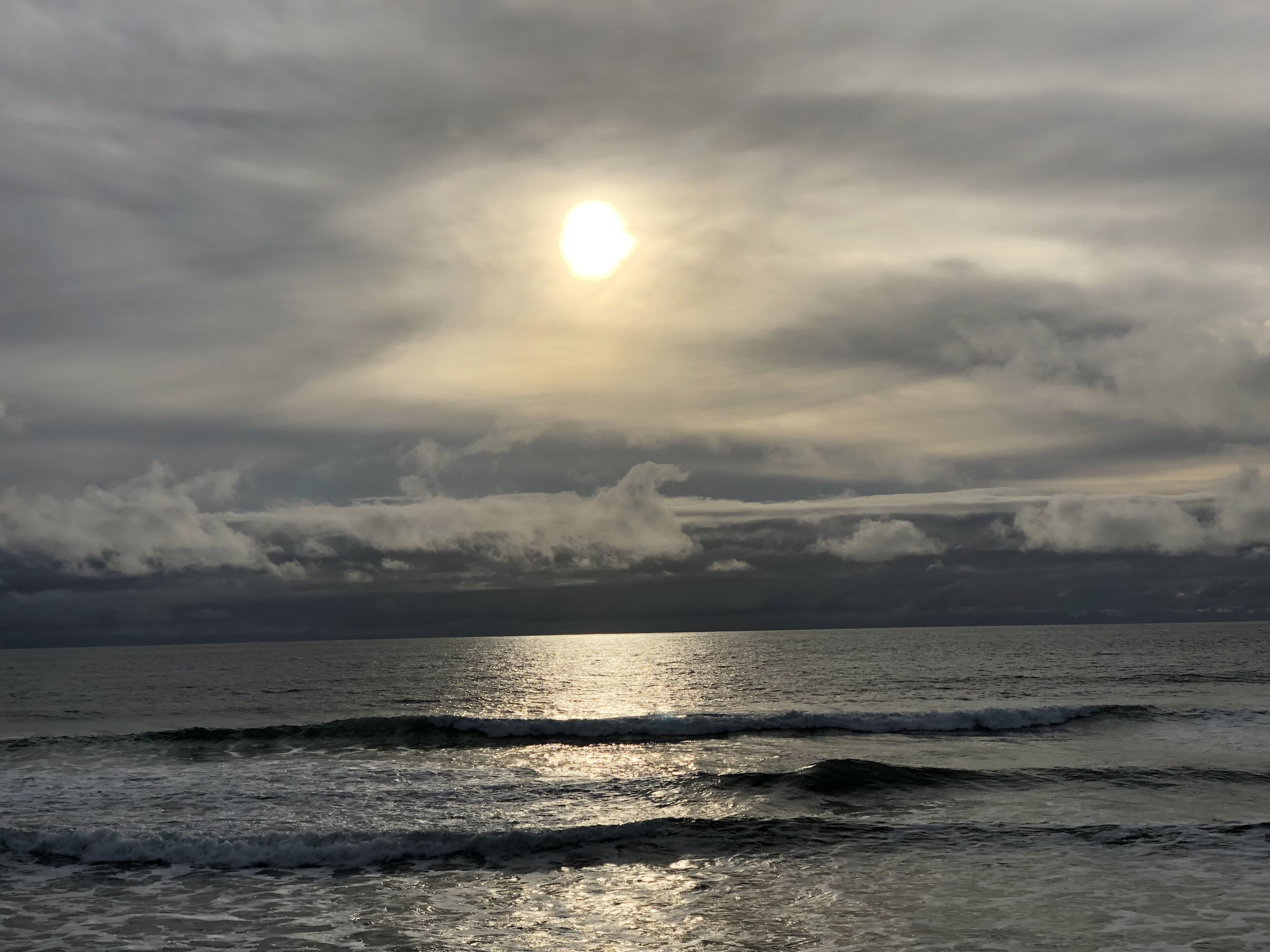 Platja amb onades i cel nuvolós, amb el sol visible.