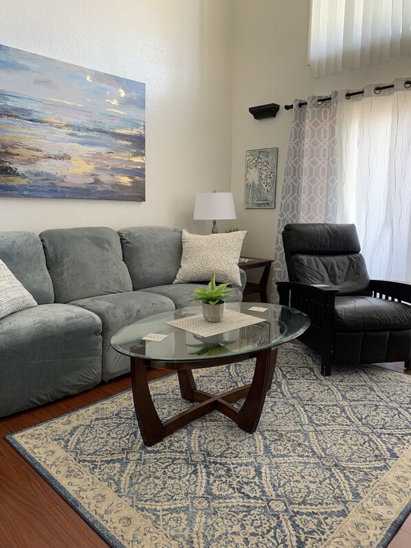Una sala de estar con un sofá gris, un sillón negro, una mesa de centro de vidrio con una planta, una lámpara y un tapete estampado.