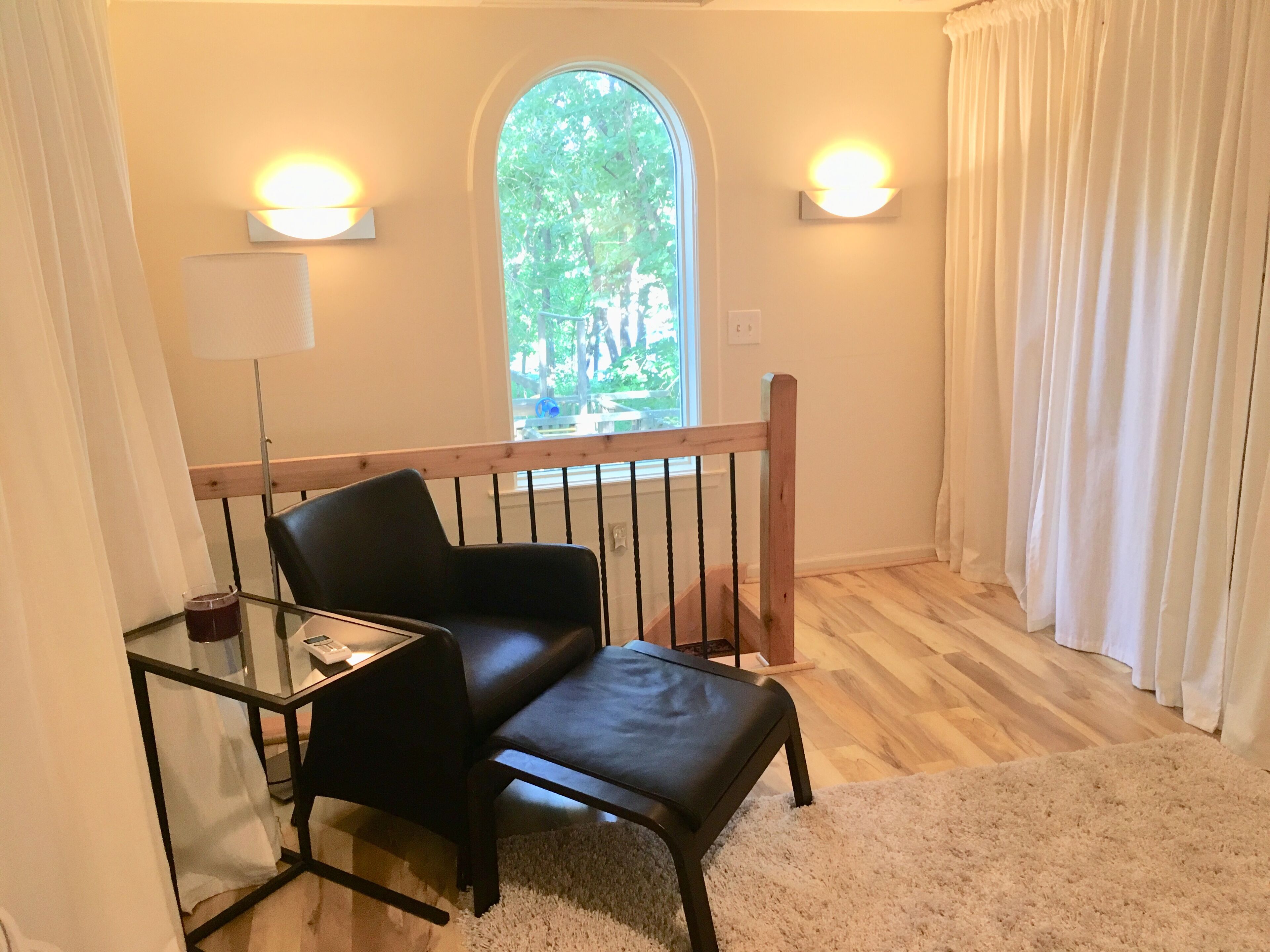 A room with a black leather chair, a glass table, a white rug, a wooden railing, and a window with a view of trees.