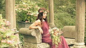 A woman in a long dress sitting on a stone bench in a garden with columns and flowering plants. - The Garden Cottage. (Stilwell)