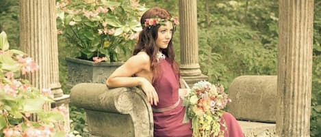 Une femme, vêtue d’une longue robe, est assise sur un banc de pierre dans un jardin orné de colonnes et de plantes en fleurs.