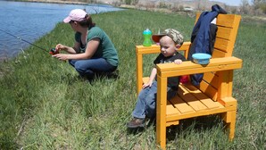 Una mujer pescando junto a un lago, con un niño sentado en una silla de madera cerca.