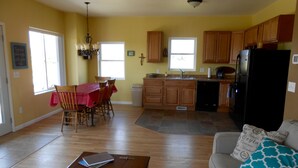 Amplia sala de estar con sofá y mesa de centro, comedor con mesa y sillas rojas, y cocina con armarios de madera y refrigerador negro.