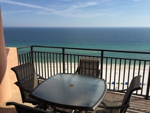Een balkon met een glazen tafel en twee stoelen, met uitzicht op een strand en de oceaan.