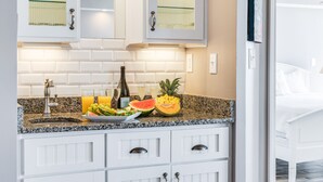 A modern kitchen with white cabinets, a granite countertop, and a variety of fruits and a bottle of wine. - Stay @ Villa Carolina on Folly Beach (Folly Beach)
