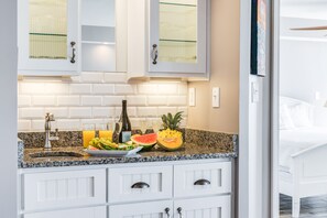 A modern kitchen with white cabinets, a granite countertop, and a variety of fruits and a bottle of wine. - Stay @ Villa Carolina on Folly Beach (Folly Beach)
