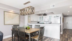A dining area with a wooden table and chairs, a hanging light fixture, and a kitchen with white cabinets and appliances in the background. - Stay @ Villa Carolina on Folly Beach (Folly Beach)