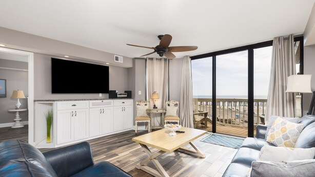 A modern living room with a flat-screen TV, a ceiling fan, a dining area with two chairs, and a balcony with a view of the ocean. - Stay @ Villa Carolina on Folly Beach (Folly Beach)