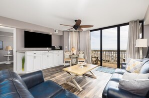 Uma sala de estar moderna com televisão de tela plana, ventilador de teto, área de jantar com duas cadeiras e varanda com vista para o oceano.