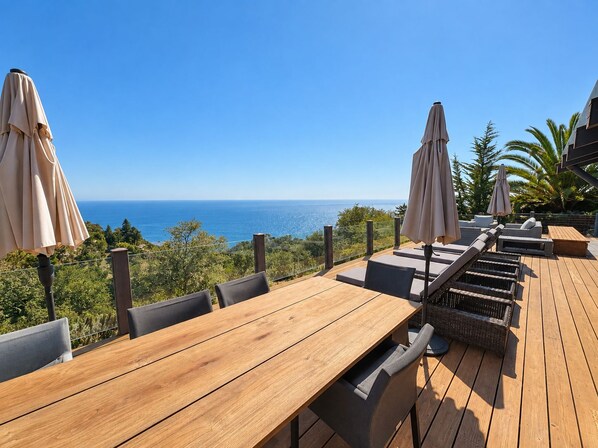 Een terras met houten tafels en stoelen, twee parasols en uitzicht op de oceaan.