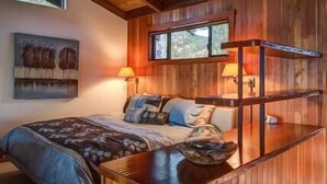 A wooden cabin-style bedroom with a bed, two bedside lamps, a window, and a painting on the wall. - Central Big Sur Retreat - Pool, Spa, Guest House + 5 Acres (Big Sur)