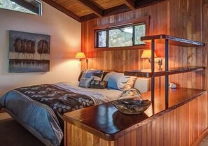 A wooden cabin-style bedroom with a bed, two bedside lamps, a window, and a painting on the wall.