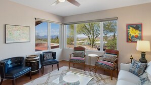 Amplia sala de estar con ventilador de techo, ventanas grandes con vistas al desierto y diversas opciones de asientos, incluyendo un sofá y sillones.