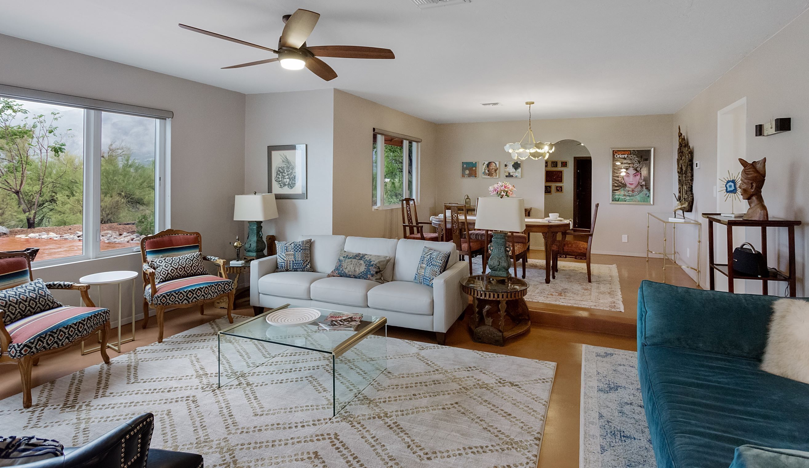 A spacious living room with a white sofa, patterned armchairs, a glass coffee table, and a dining area with a chandelier.