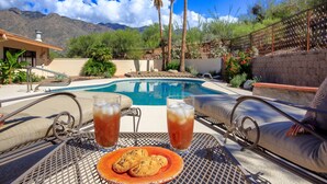 Zona junto a la piscina con vistas a montañas, una alberca y una mesa con bebidas y galletas.