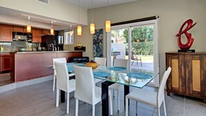 Une cuisine moderne avec un coin repas, comprenant une table à plateau en verre, des chaises blanches et des luminaires suspendus.
