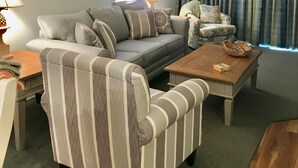 A living room with a sofa, armchair, and coffee table, a window with blinds, a lamp, and a painting on the wall. - End Unit:  8th Floor Ocean Front (Myrtle Beach)