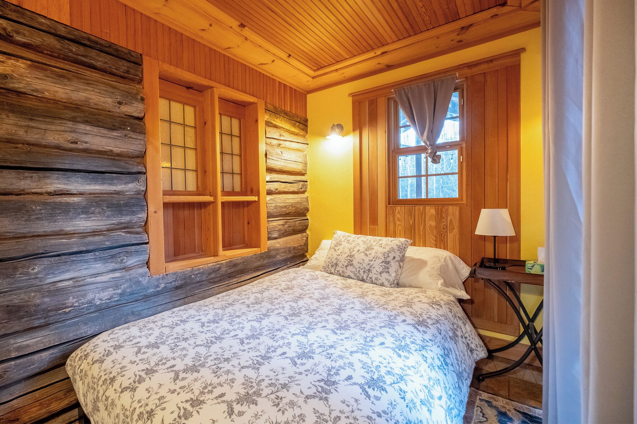 A log cabin bedroom with a bed, wooden walls, a window with curtains, and a nightstand with a lamp.