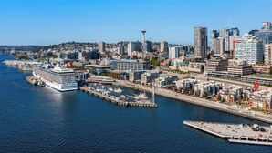 Un paisaje urbano frente al agua con edificios modernos, una torre destacada y un crucero amarrado en un puerto deportivo.