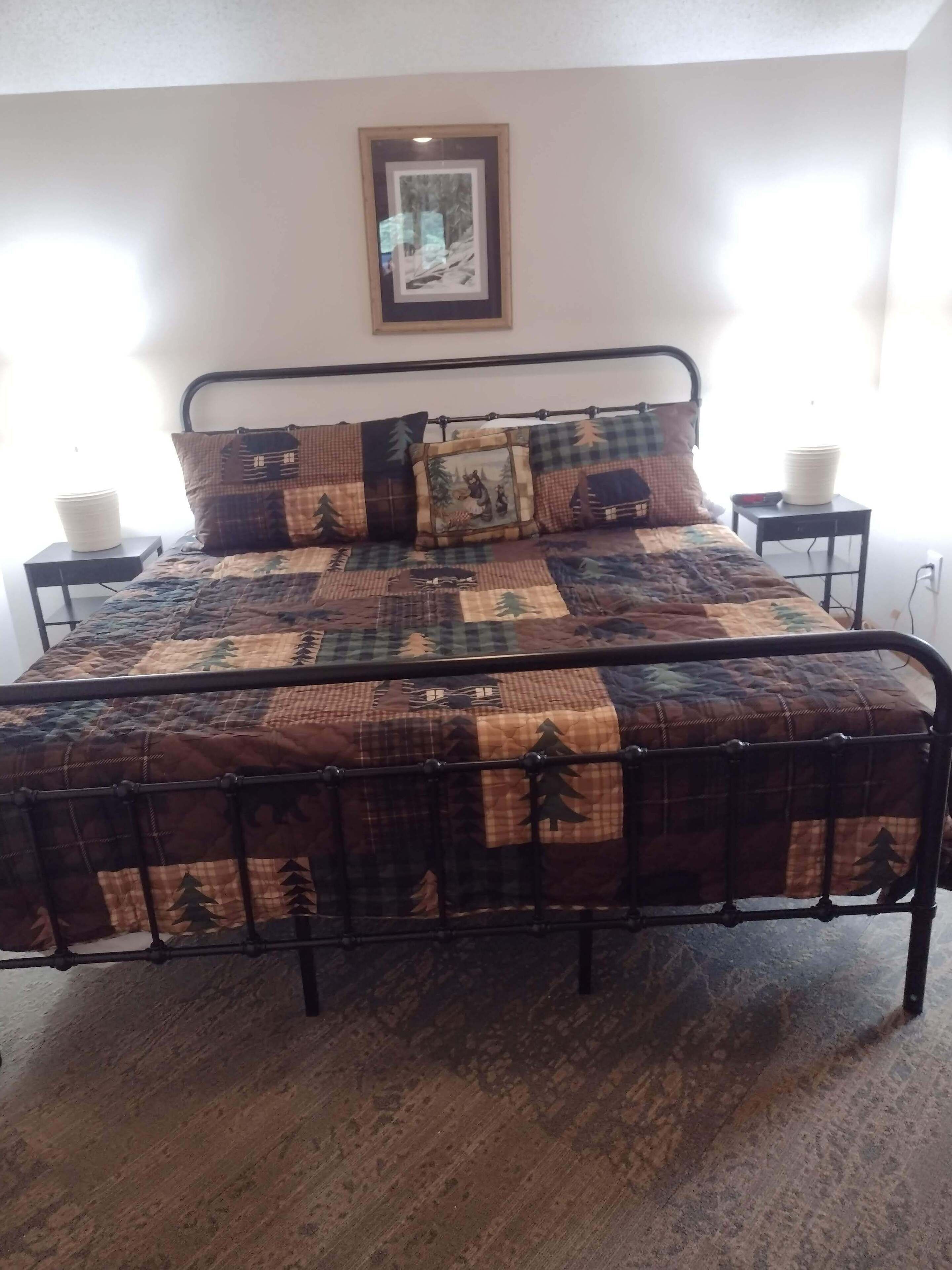 A bed with a quilted bedspread featuring a forest pattern, flanked by two bedside tables with lamps, a framed picture on the wall, and a window providing natural light.