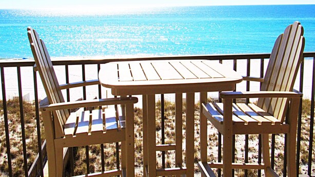 A wooden deck with two Adirondack chairs and a small table, overlooking a beach with clear blue water.