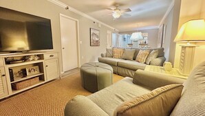 A living room with a large flat-screen TV, a sofa set, a ceiling fan, and a lamp.