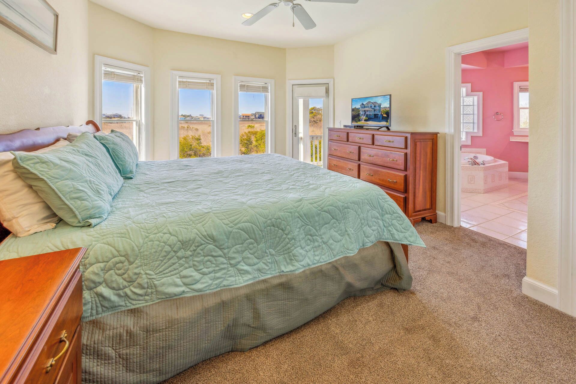 A bedroom with a large bed, a dresser, and a vanity with a mirror. There are multiple windows and a door leading to another room.