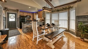 A modern kitchen with wooden cabinets, a central island, and a dining area with a bench and table. - Family Getaway Close to Parkway-w/ Theater and Game Room (Pigeon Forge)