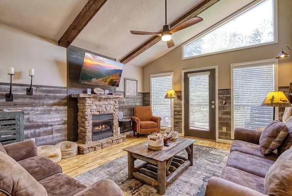 A cozy living room with a fireplace, comfortable seating, and a large window with blinds.