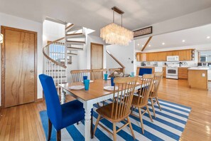 A modern kitchen with wooden cabinets, a central island, and a dining area with a striped rug and blue chairs. - Oceanfront Home in Waves with Hot Tub, Grill, and Screened Porch (Waves)