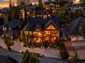 A multi-story log cabin with a central deck and multiple windows, surrounded by trees and a driveway. - 🐻🏰 The Big Bear Castle | Luxury 6,000 Sq Ft • 6BR/7BA • Hot Tub • Lakeview! (Big Bear)