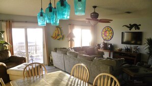 A living room with a sofa, a dining table, and chairs. There is a television and a decorative wall hanging.