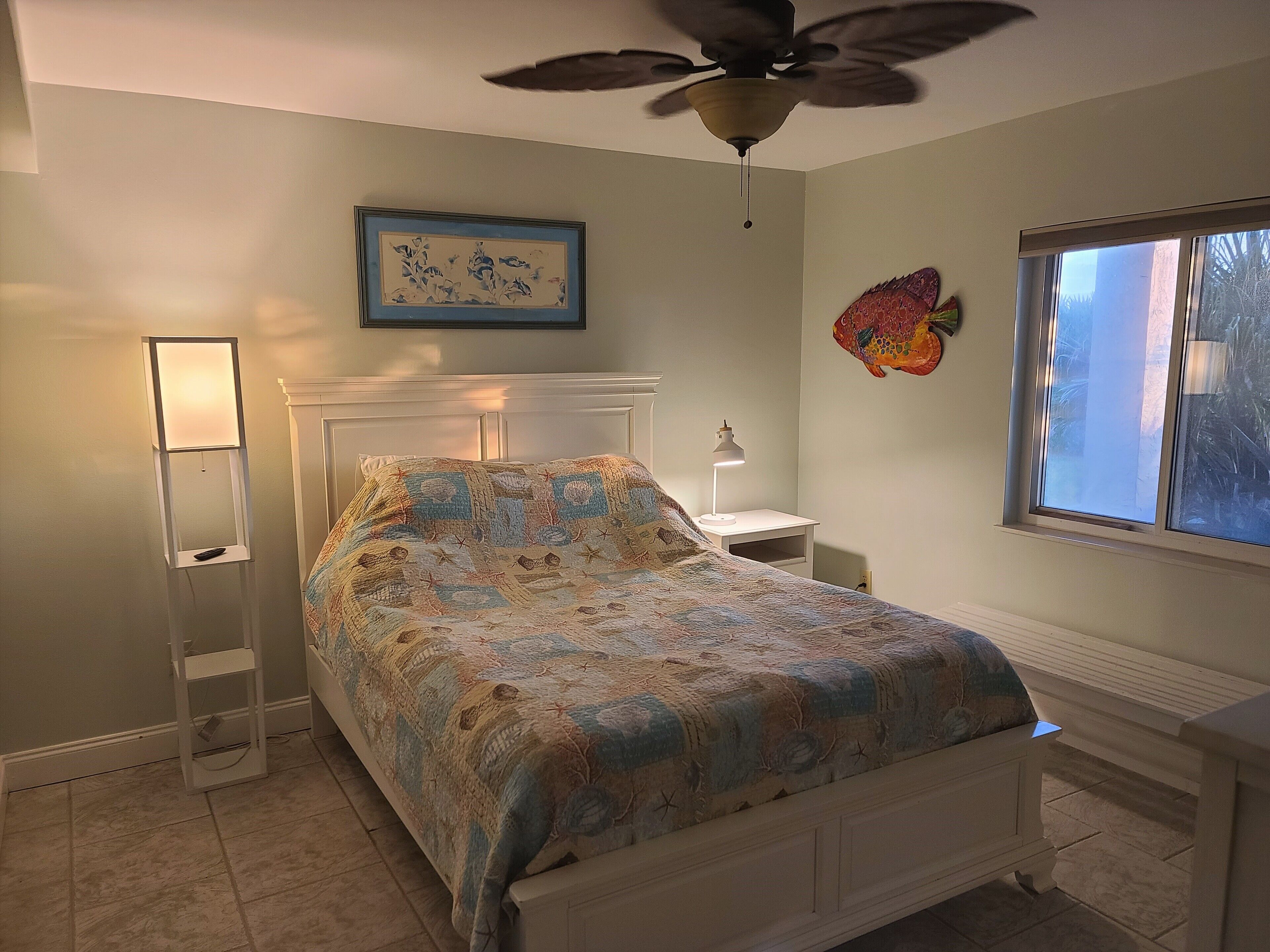 Un dormitorio con cama, ventilador de techo, mesita de noche con lámpara, un cuadro en la pared y una ventana con vistas.