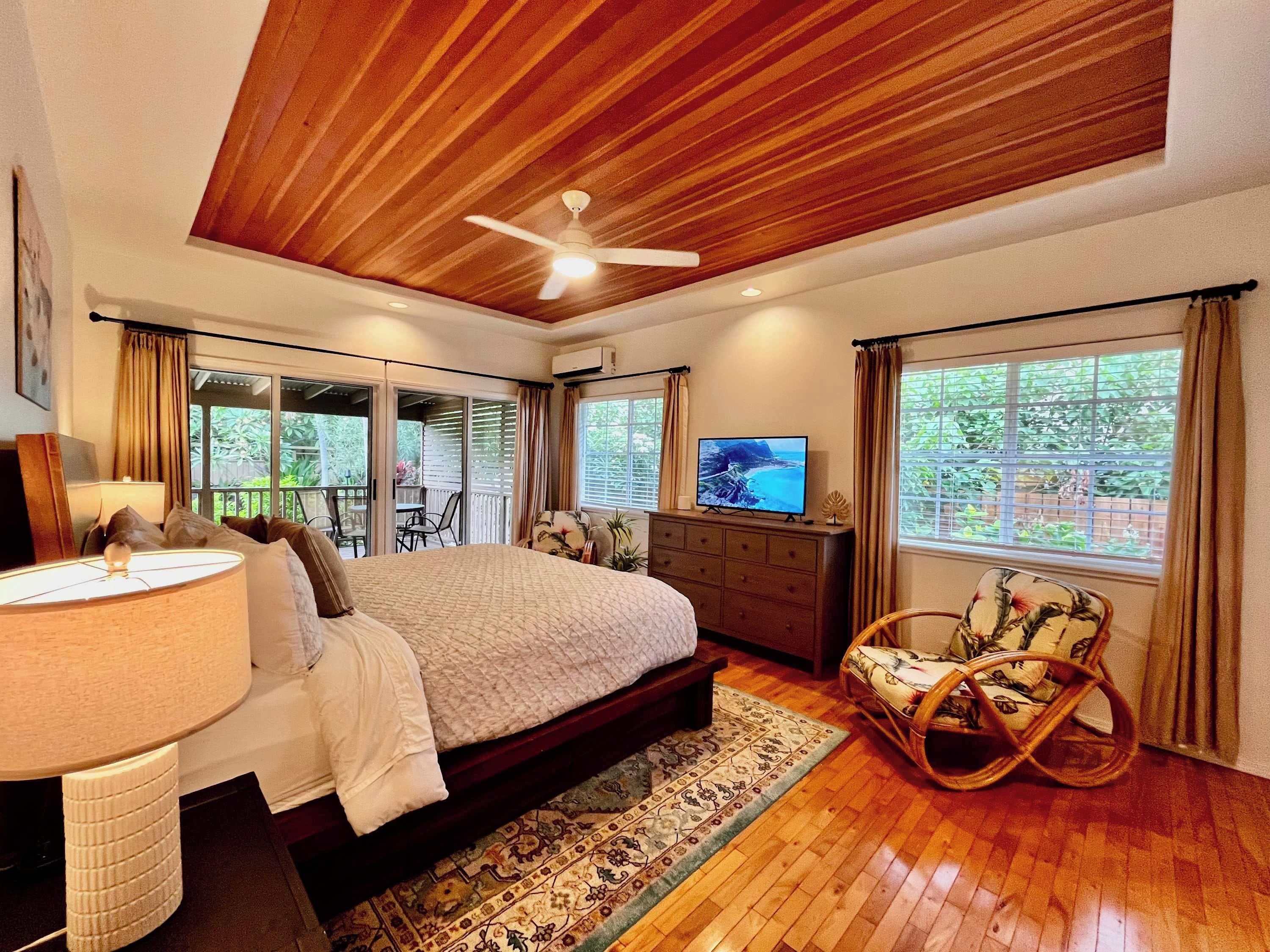 A bedroom with a bed, a television, a desk with a lamp, a rocking chair, and a view of the outdoors.