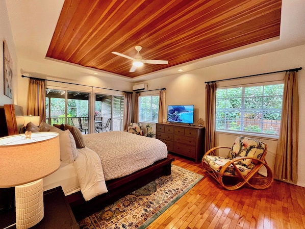 A bedroom with a bed, a television, a desk with a lamp, a rocking chair, and a view of the outdoors.
