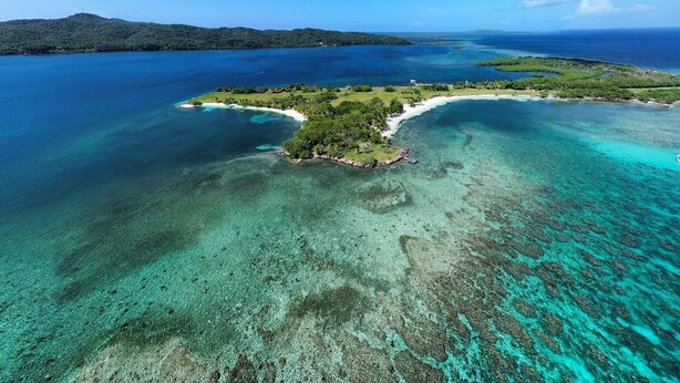 Vue aérienne d’une île tropicale aux eaux turquoise et limpides, entourée d’une végétation luxuriante et dominée par une chaîne de montagnes lointaine.