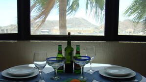 A dining table set with plates, glasses, and a bottle of wine, with a view of palm trees and mountains through a window. - Good Value Cabo Condo with Pool at Marina Sol. Walk to Beach, Bars!! (Cabo San Lucas)