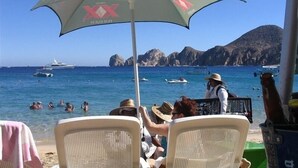 A beach scene with a large umbrella labeled 'XX Dos Equis' and a view of the sea with boats and people swimming. - Good Value Cabo Condo with Pool at Marina Sol. Walk to Beach, Bars!! (Cabo San Lucas)