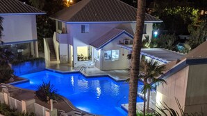 Vista nocturna de una casa con piscina, rodeada de palmeras y un patio bien iluminado.