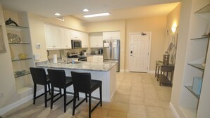 Una cocina moderna con armarios blancos, una isla central con sillas, electrodomésticos de acero inoxidable y suelo de baldosas.