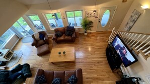 Amplia sala de estar con piso de madera, sofás de cuero, una mesa de centro y un televisor sobre un mueble bajo. Amplias ventanas ofrecen vistas al exterior.