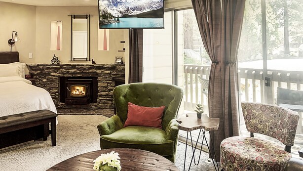 Habitación de hotel con una cama, un sillón verde, una mesita de centro redonda con un florero, un televisor y una chimenea de piedra.