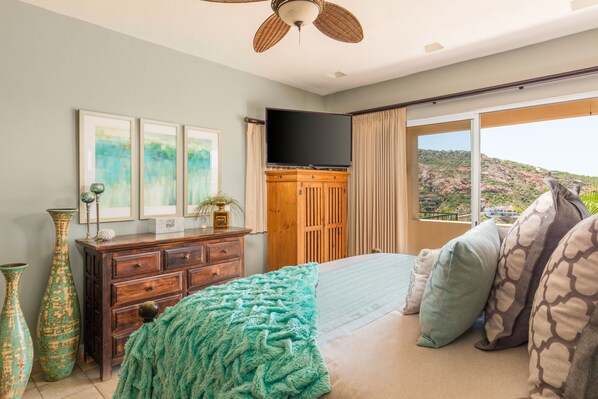 Un dormitorio con cama, cómoda, televisor de pantalla plana y ventana grande con vista a un paisaje montañoso.