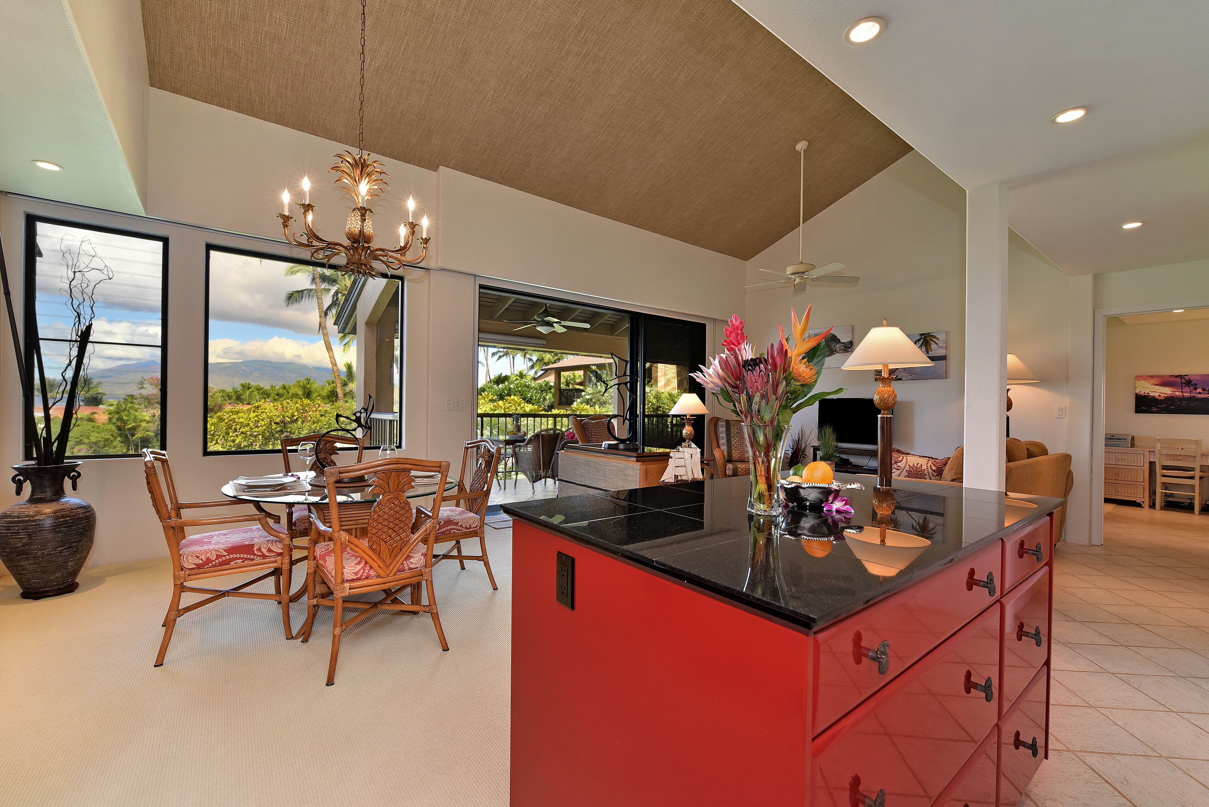 Una cocina amplia con una isla roja, zona de comedor con sillas de mimbre y ventanas grandes con vistas a la vegetación y montañas.