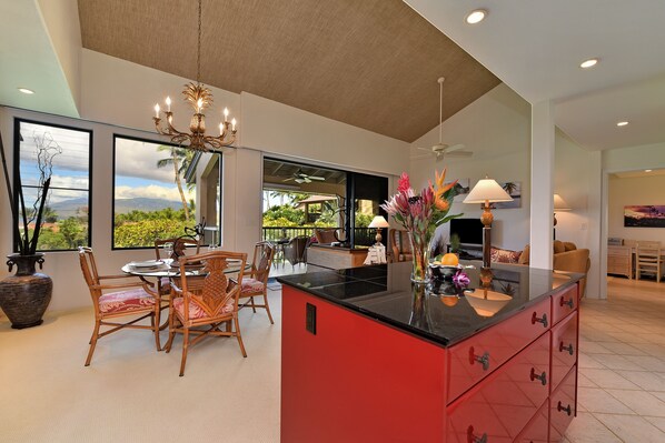A spacious kitchen with a red island, dining area with wicker chairs, and large windows offering a view of greenery and mountains.