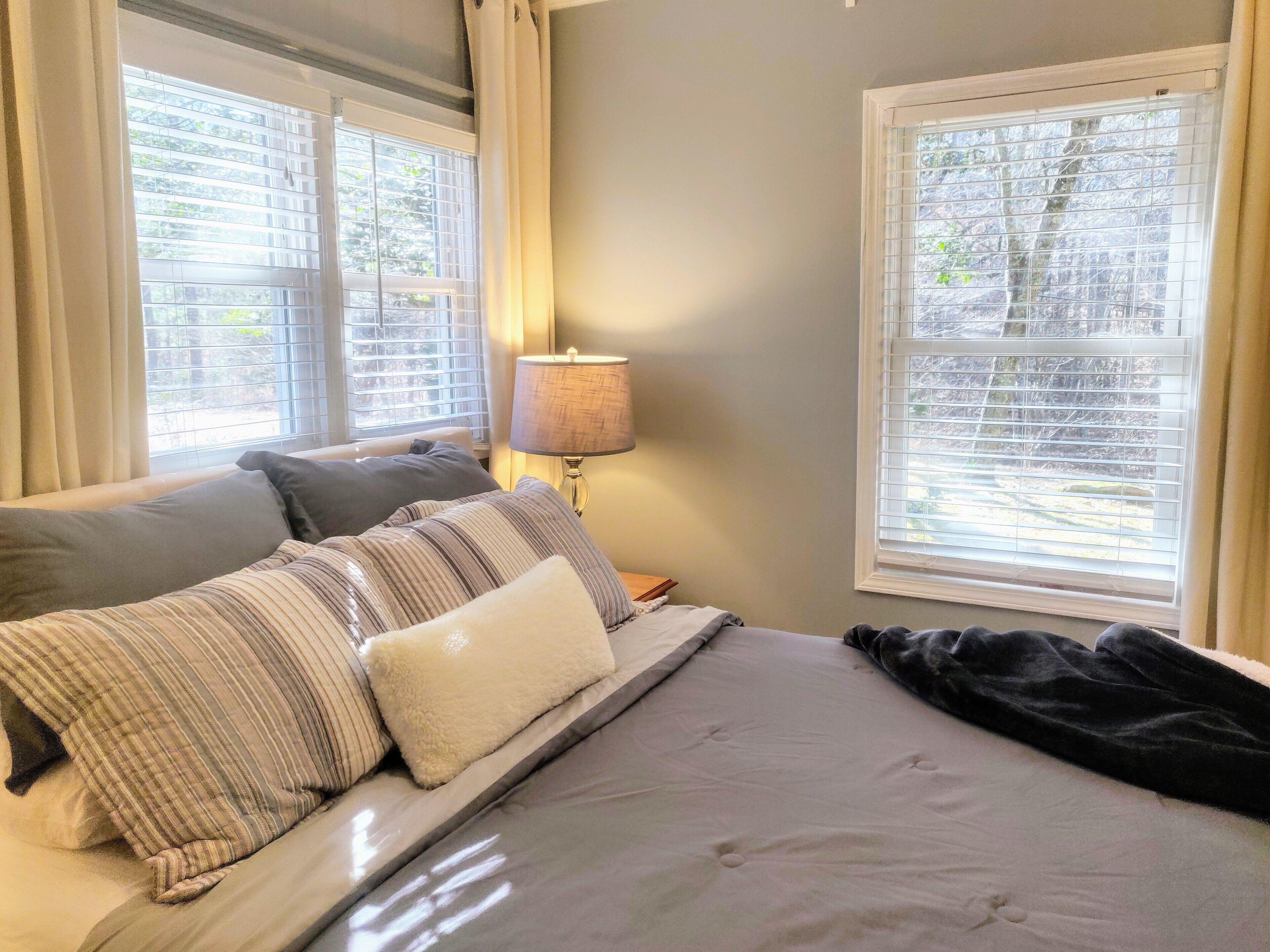 A bedroom with a bed, pillows, a lamp, and windows with blinds.