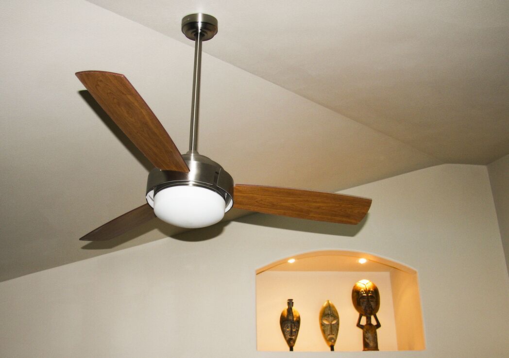 Un ventilateur de plafond avec des pales en bois et un socle métallique, dans une pièce dont le plafond est blanc et qui est dotée d’une étagère encastrée où sont exposés trois objets décoratifs.