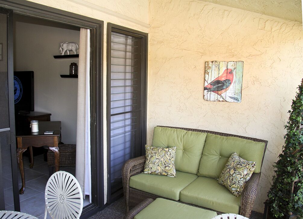 Ein grünes Sofa mit gemusterten Kissen, ein Korbstuhl und ein Wandbild mit Vogelmotiv in einem Raum mit strukturierter Wand und einer Tür zu einem weiteren Bereich.