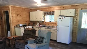 A cozy cabin-style kitchen with wooden walls, a dining table, two armchairs, a refrigerator, and a stove. - Quaint getaway to unwind and relax.  (Manistique)